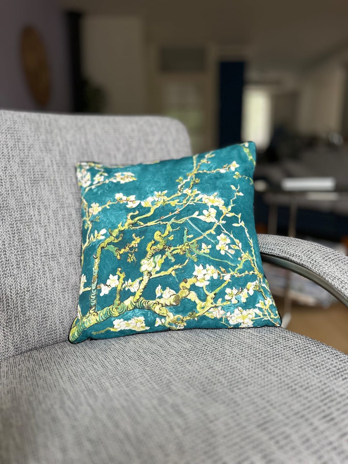 Blauwgroen kussen met wit bloemenmotief op een grijze stoel in een wazige binnenomgeving.