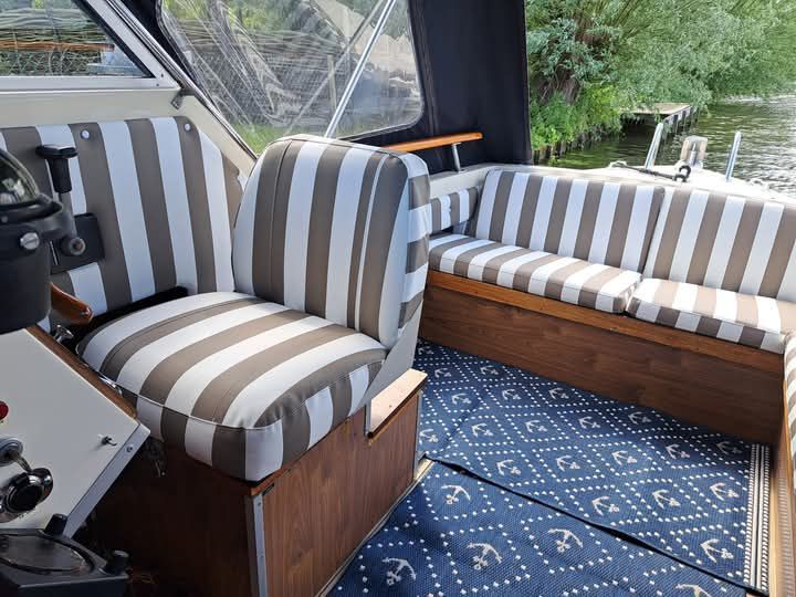 Interieur van de cockpit van een boot met gestreepte stoelen in grijs en wit, een blauw tapijt en een stuurwiel.