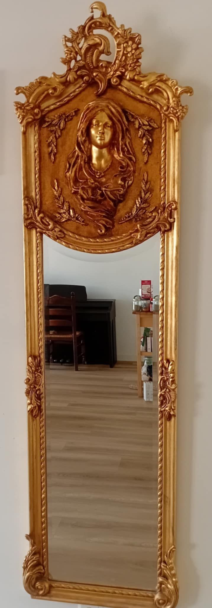 Hoge, sierlijke gouden spiegel met een decoratief borstbeeld bovenaan, dat een kamer met meubels en voorwerpen weerspiegelt.