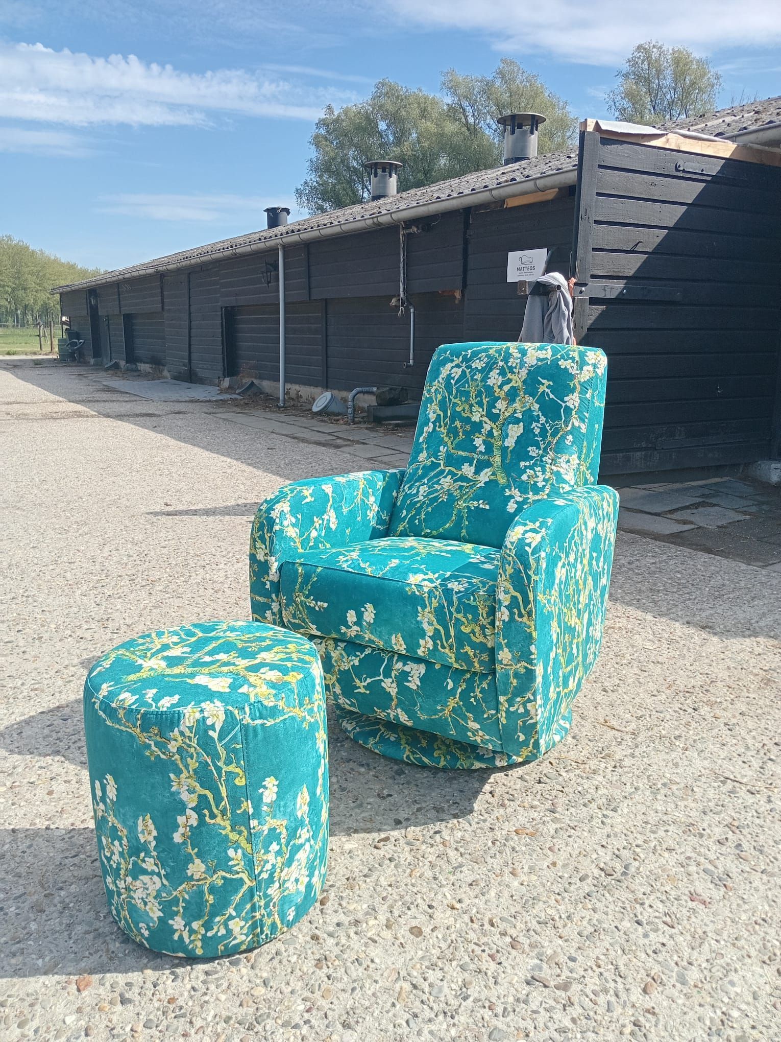 Buiten staat een turquoise bloemenfauteuil en voetenbankje; op de achtergrond het lange, donkere gebouw.