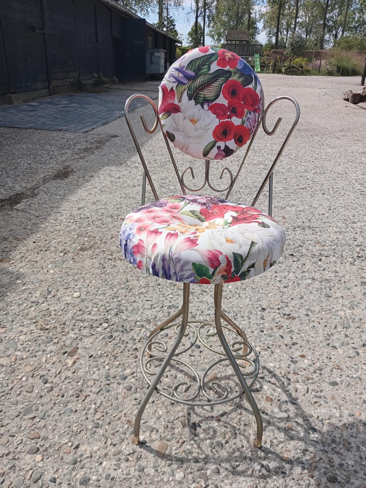 Speelse metalen kruk met bloemenprint op de zitting en rugleuning voor buiten op grind.