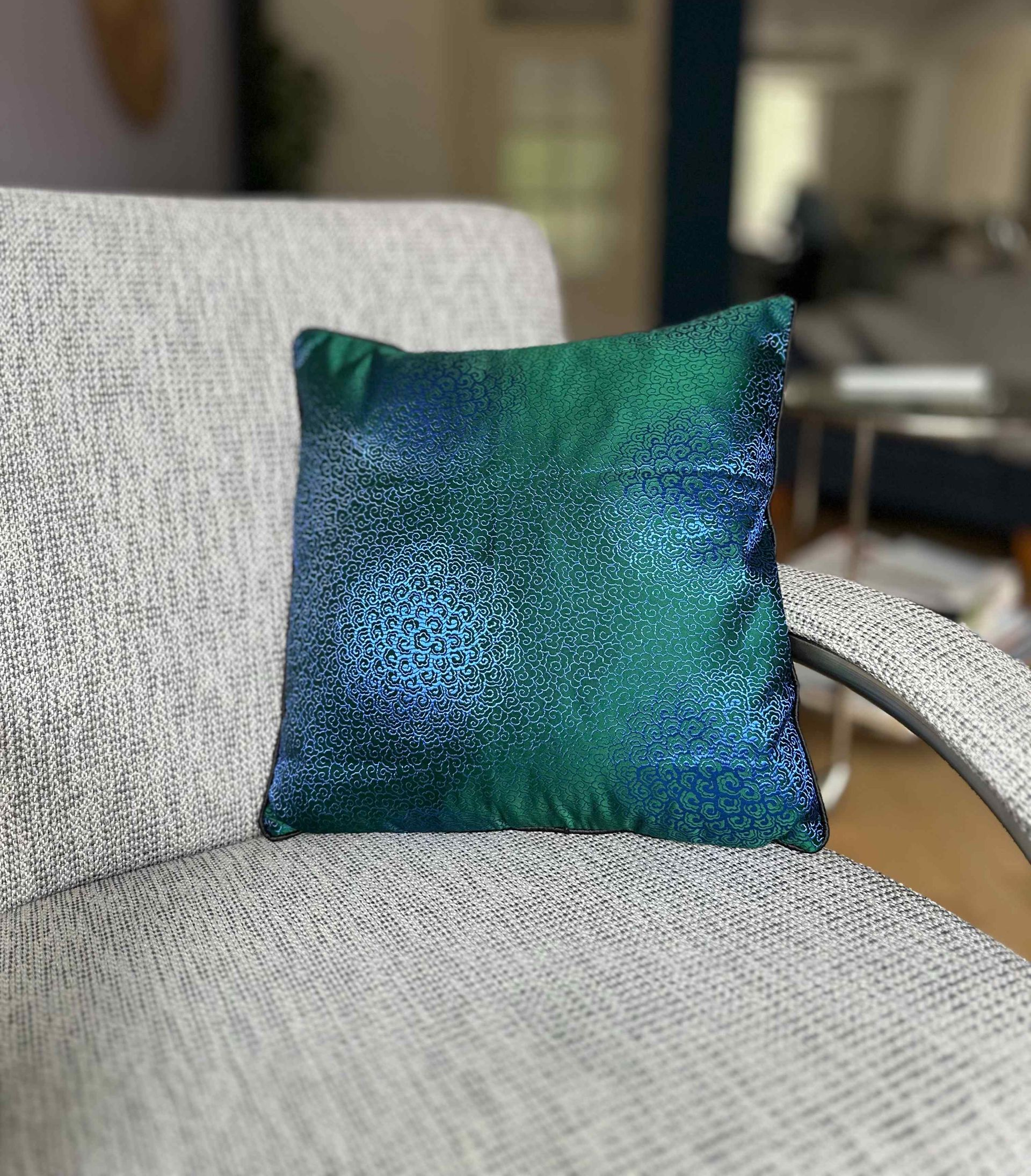 Vierkant sierkussen met een abstract blauw en groen ontwerp ligt op een grijze stoel.