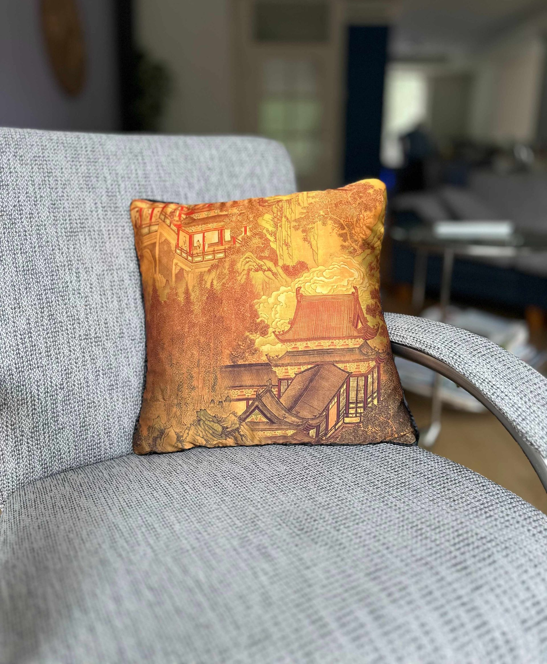 Oranje bedrukt kussen op een grijze stoel met patroon; op de achtergrond het interieur van de kamer.