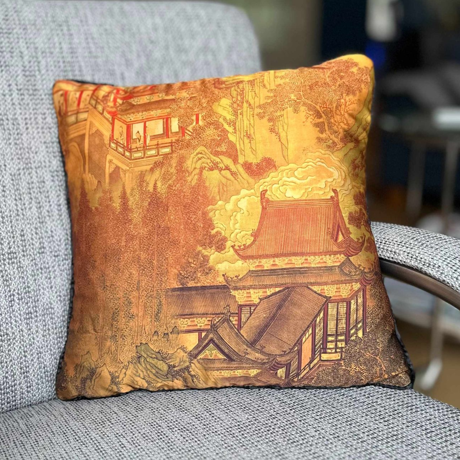 Decoratief kussen met een Oost-Aziatisch landschap in goudtinten, rustend op een grijze fauteuil.