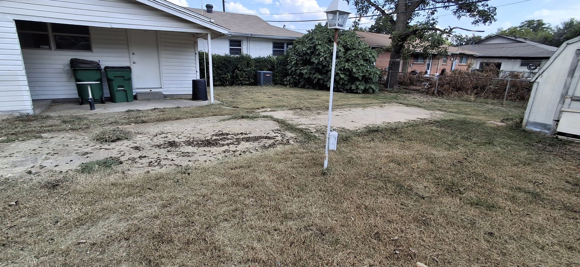 Backyard with patchy lawn, white house with green trash cans, a light pole, and shrubbery.