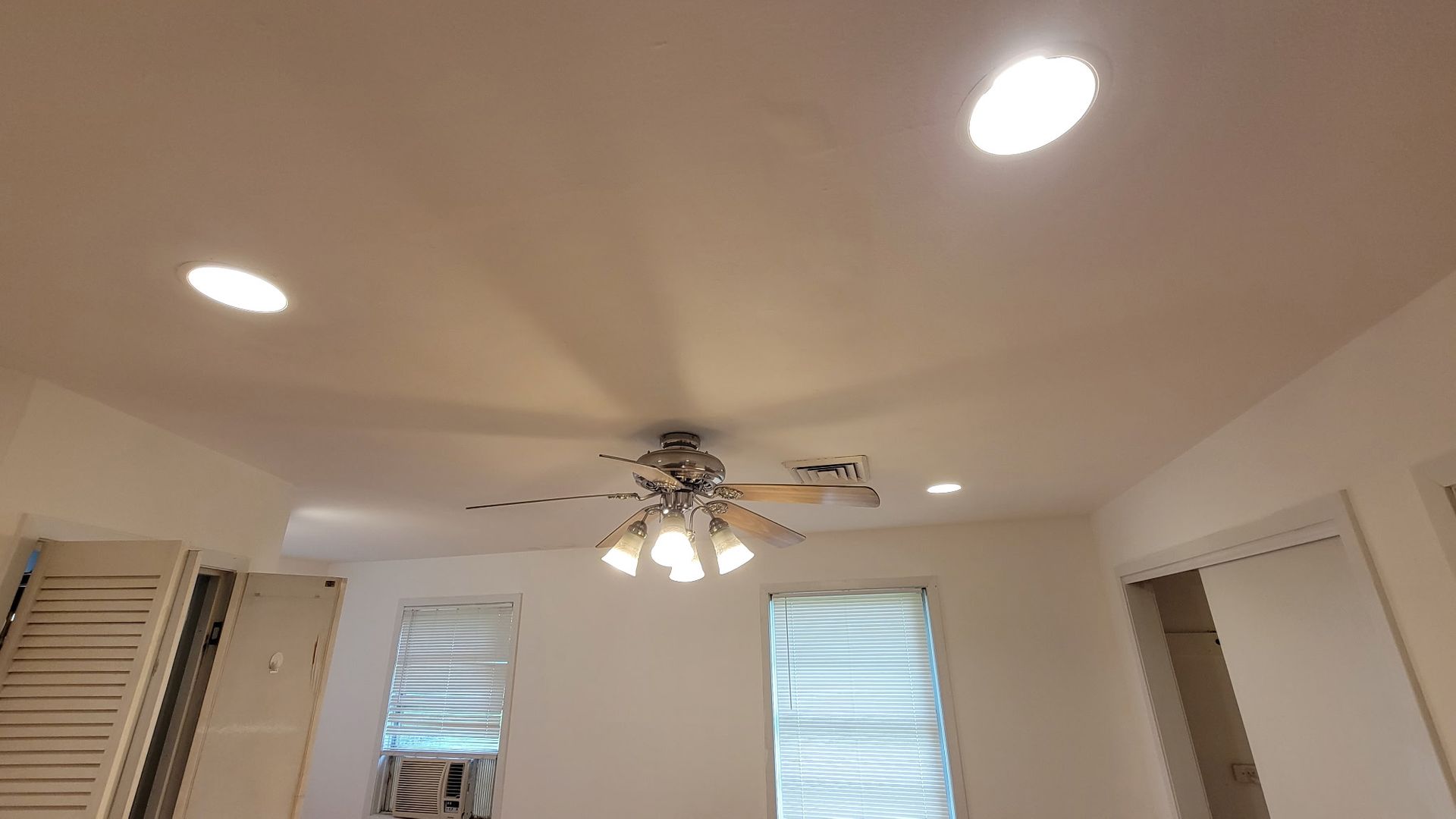 A room ceiling with a ceiling fan, recessed lights, and two windows with blinds.