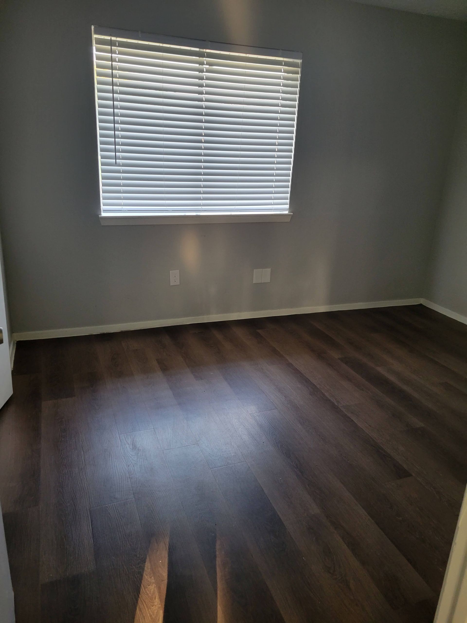 Empty room with dark wood-look flooring, gray walls, and a window with blinds.