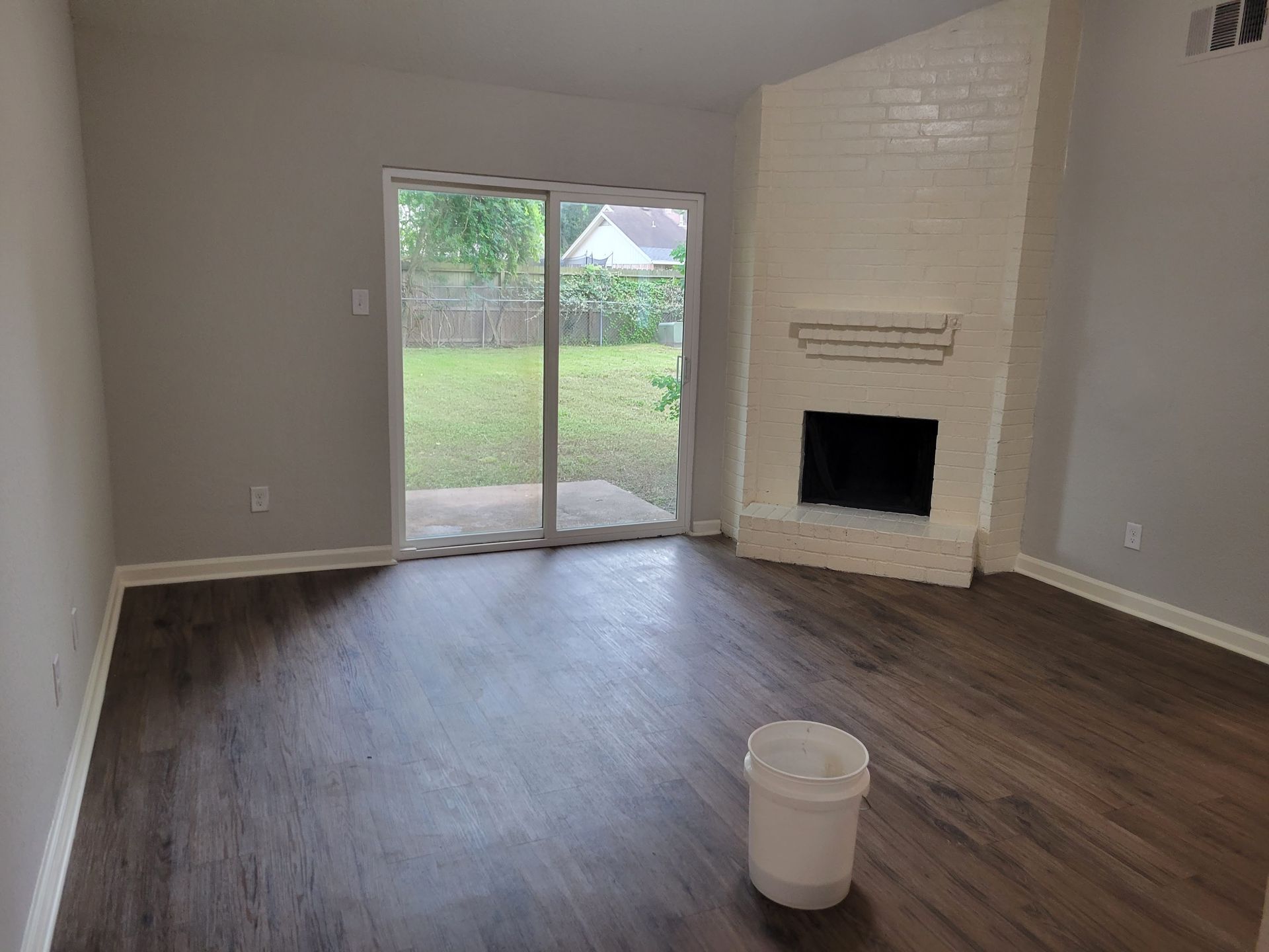 Empty living room with dark wood-look flooring, sliding glass door to backyard, white painted brick fireplace.