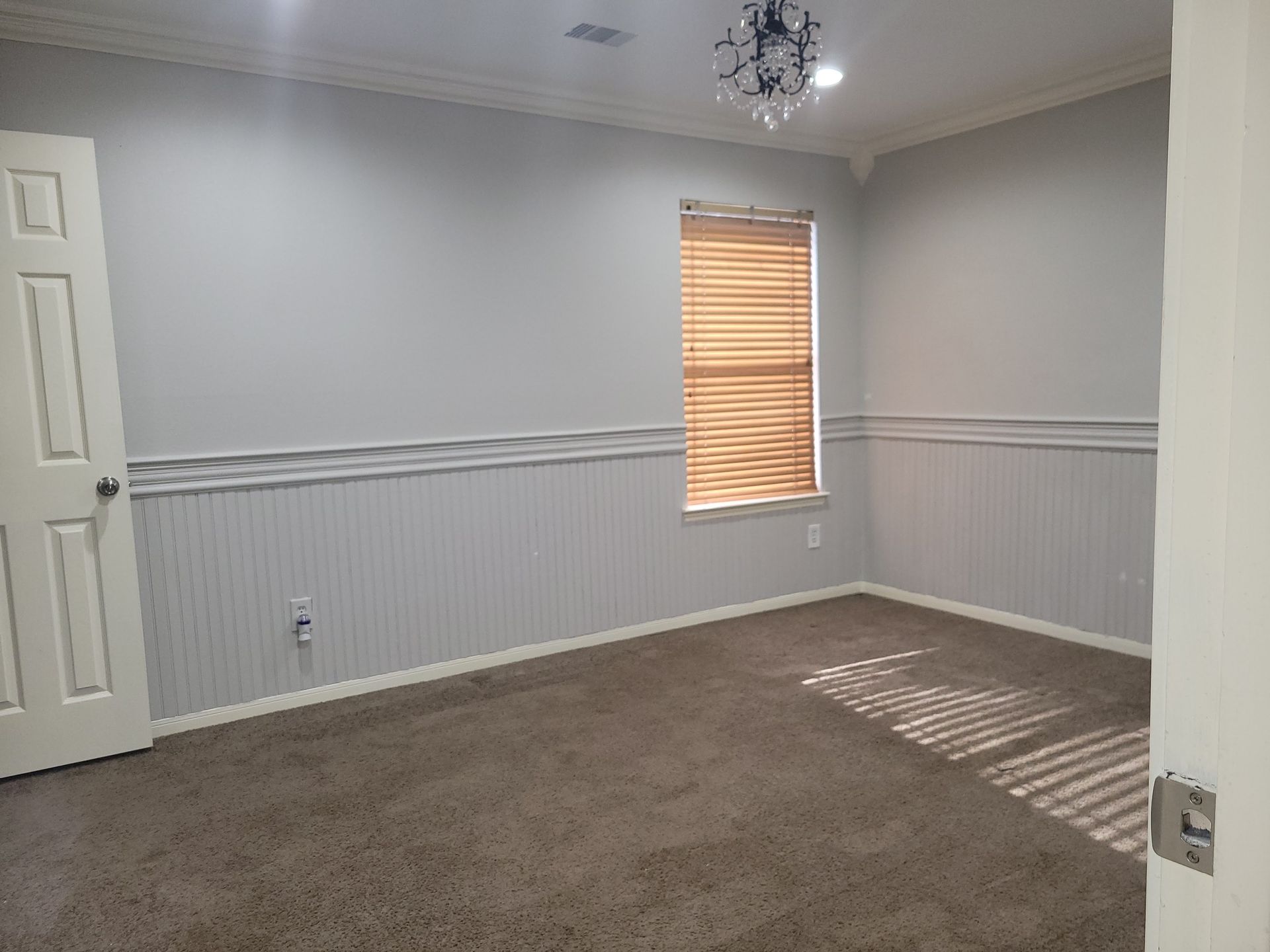 Empty room with light gray walls, wood trim, brown carpet, window with blinds, and white door.