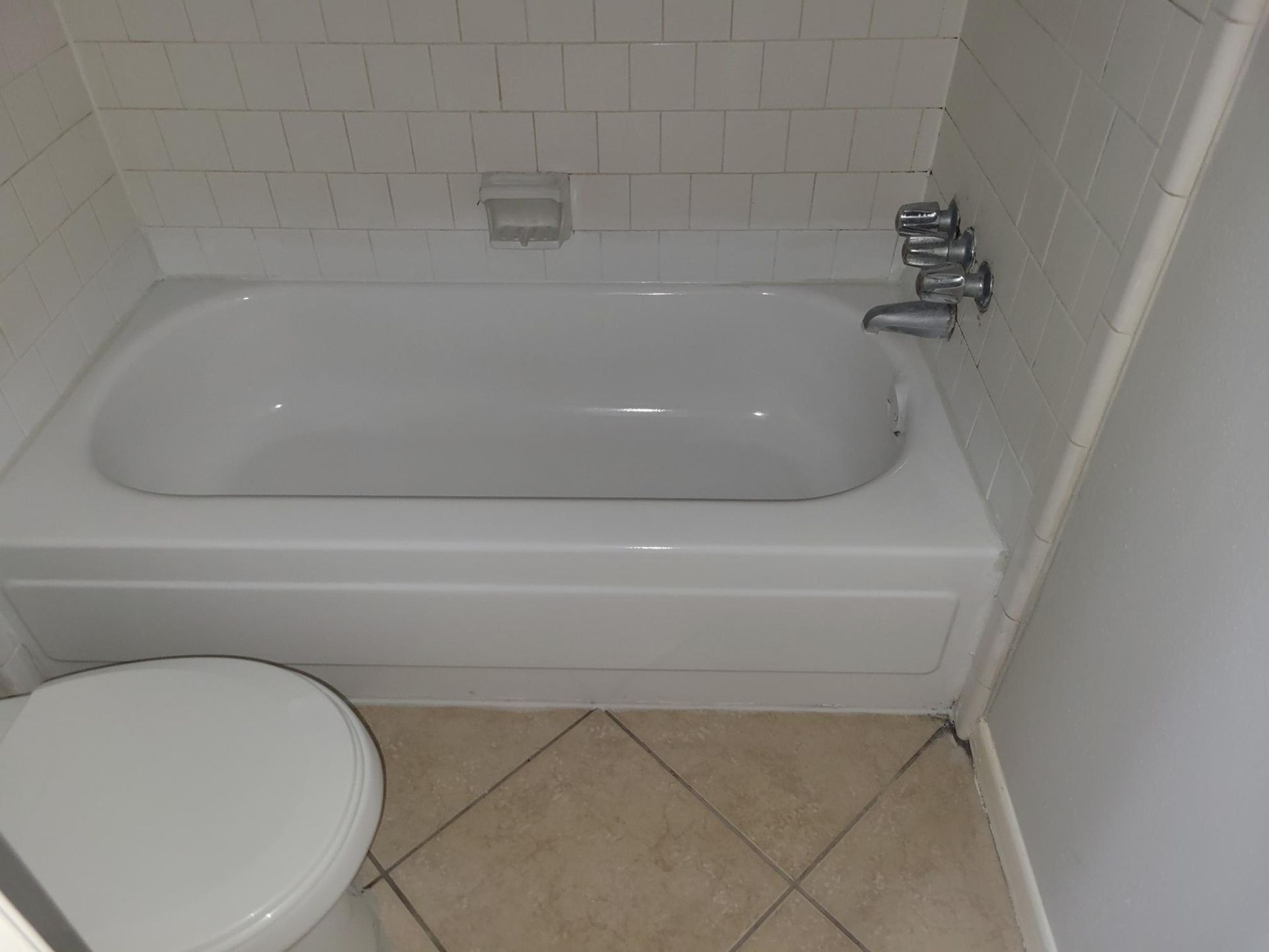 White bathtub with a toilet in a bathroom, beige tiled floor and white wall.