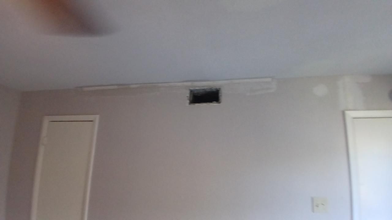 A room interior with an air vent in the wall, flanked by doors, with a partial view of a ceiling fan.