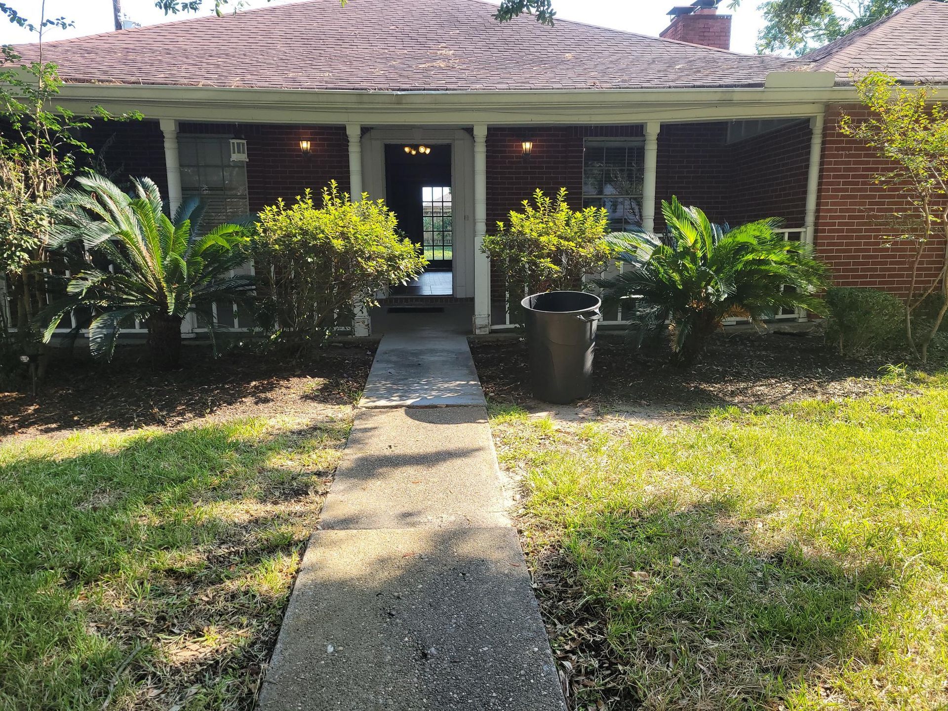 Brick house exterior with a walkway, porch, bushes, and palm trees.