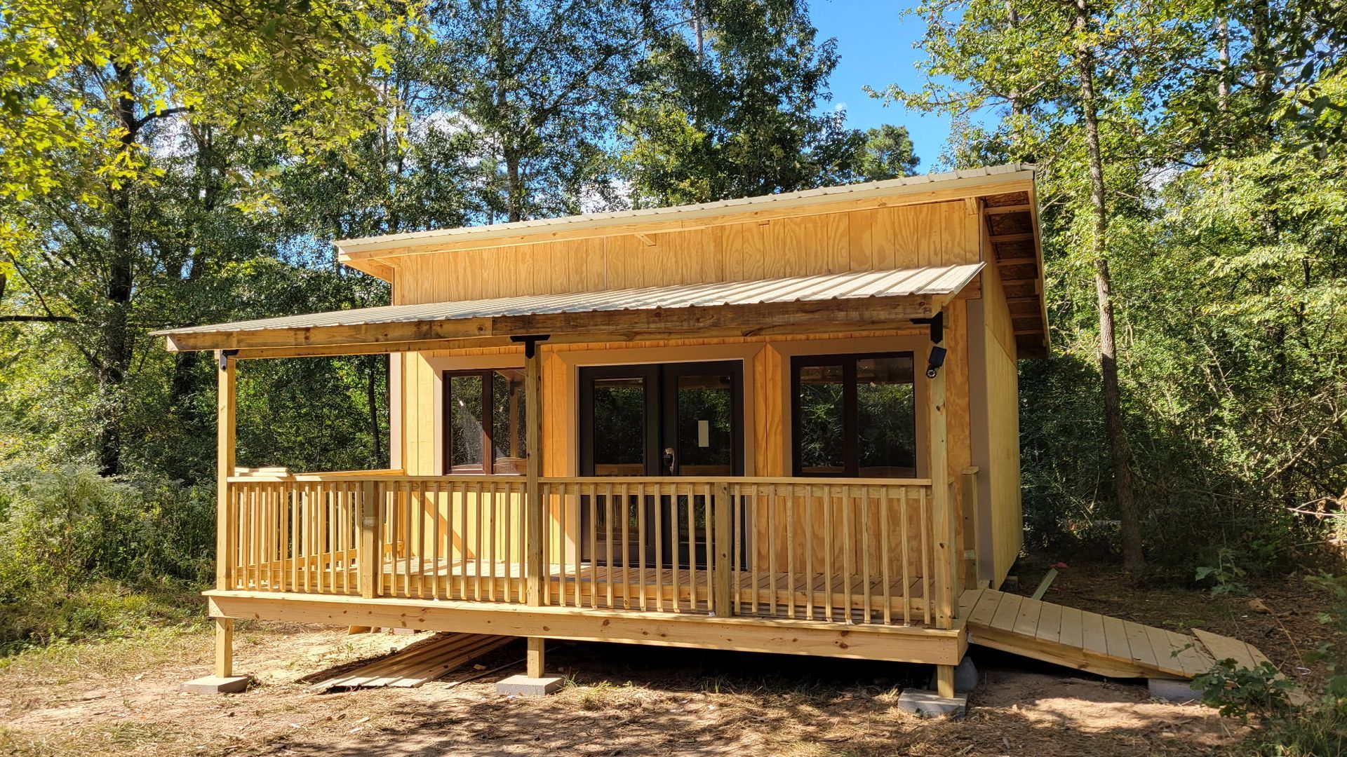 Wooden cabin with porch and ramp, surrounded by trees under a blue sky.