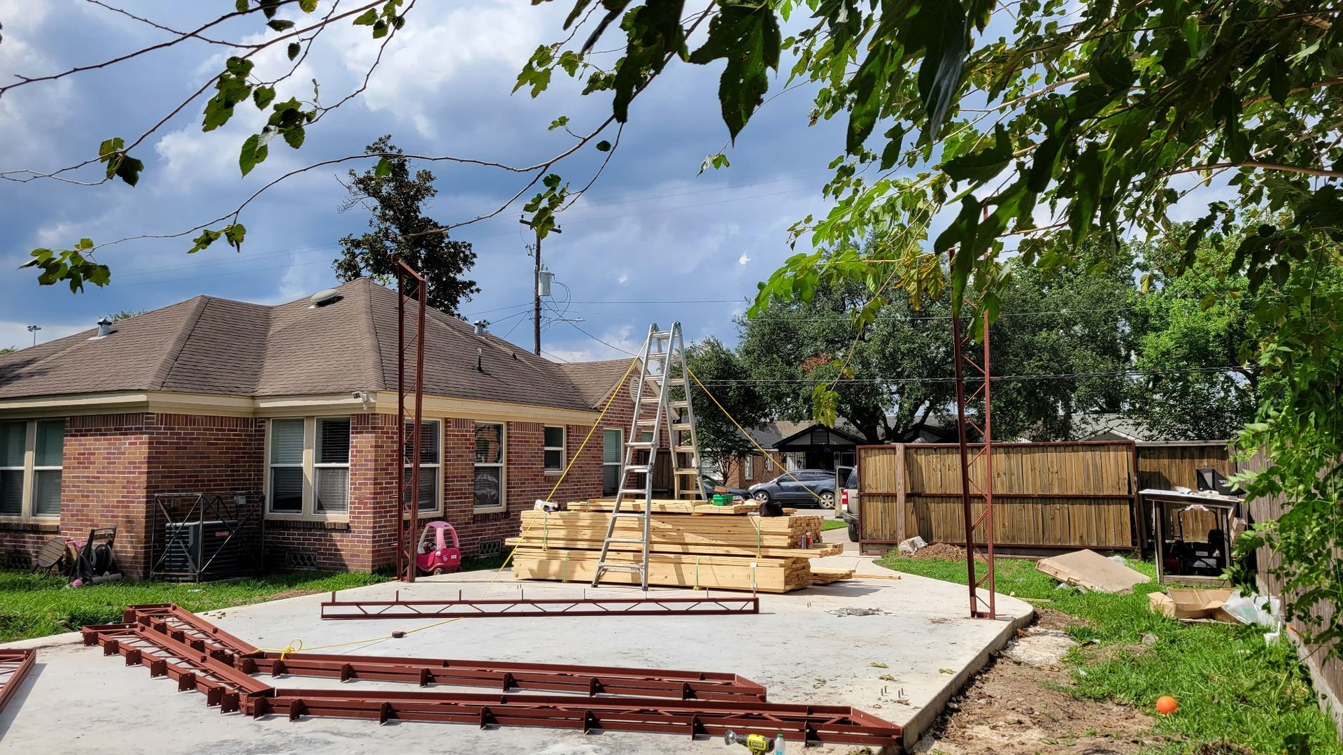 Backyard construction site; concrete slab, lumber, ladders, brown fence, brick house, cloudy sky.
