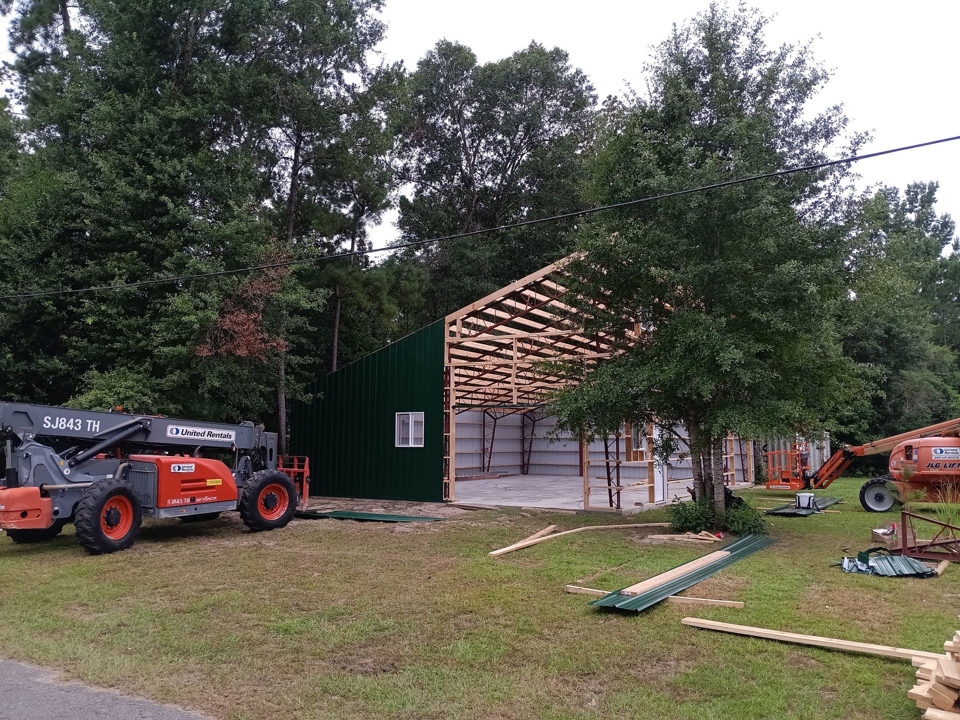 Construction site with green metal siding building under construction; orange machinery and tools in yard.