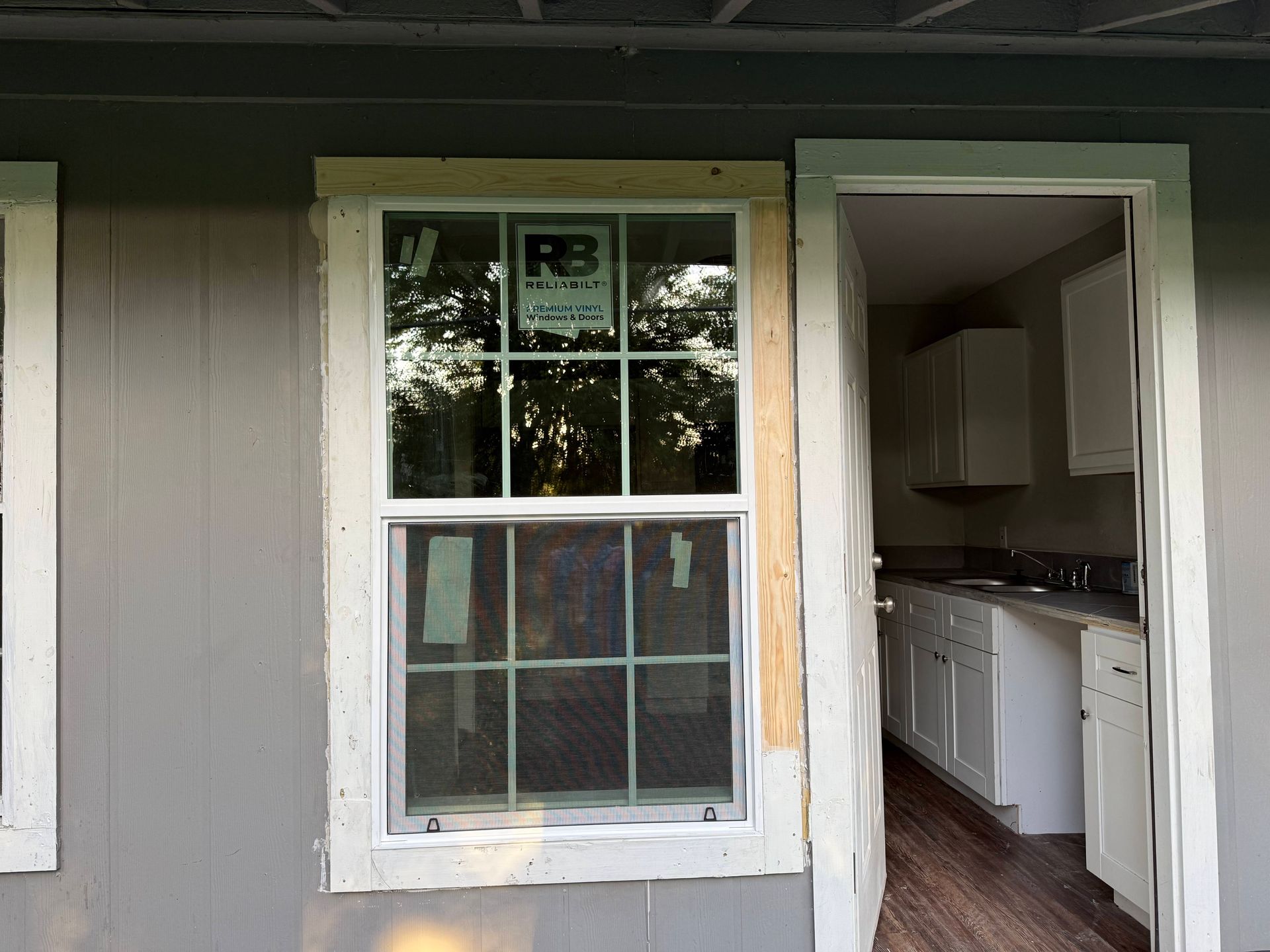 Window and doorway in a gray building. A white-framed window is next to an open doorway leading to a kitchen.