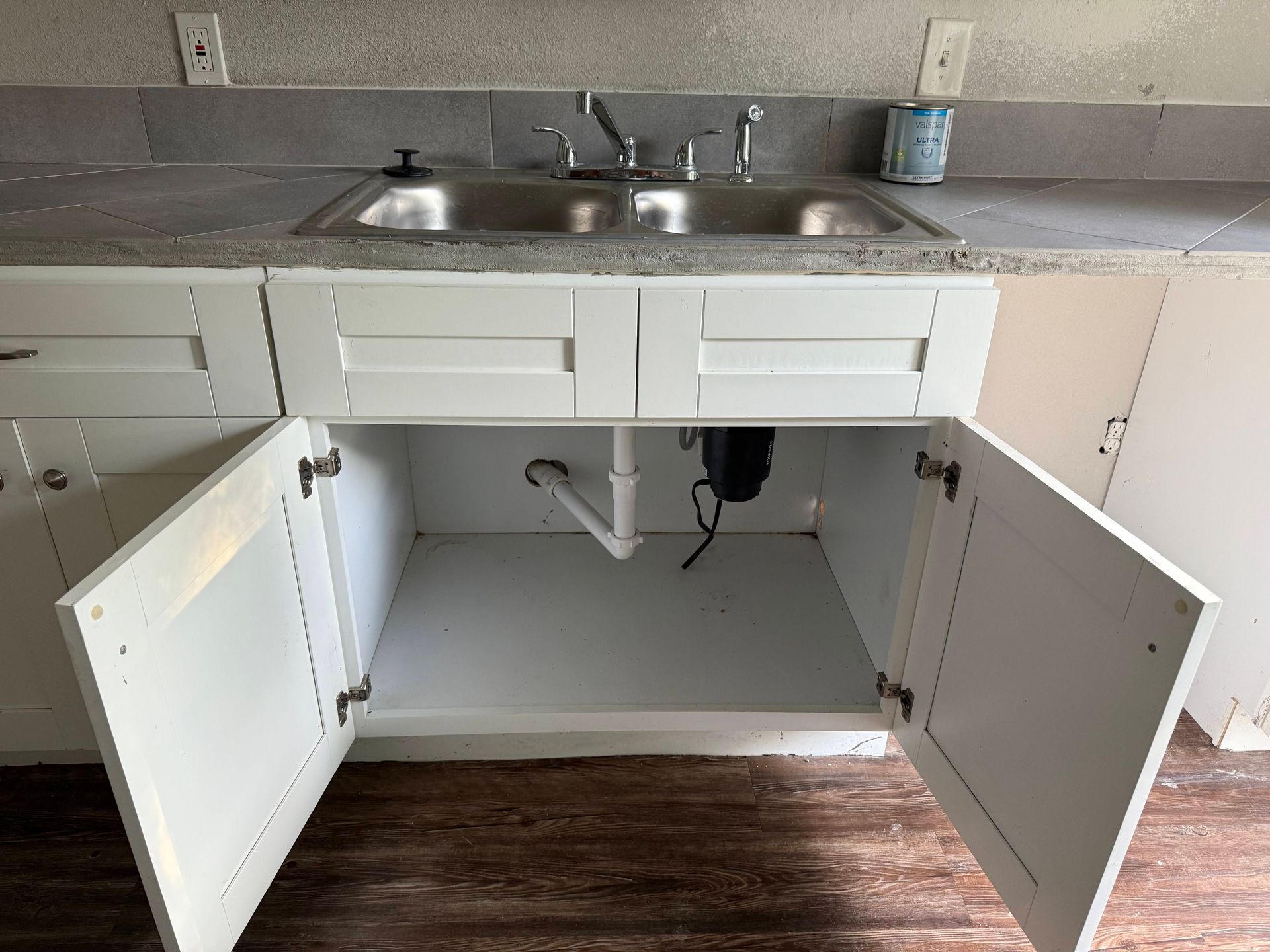 Kitchen sink with open white cabinet doors, showing pipes and a disposal under the sink.
