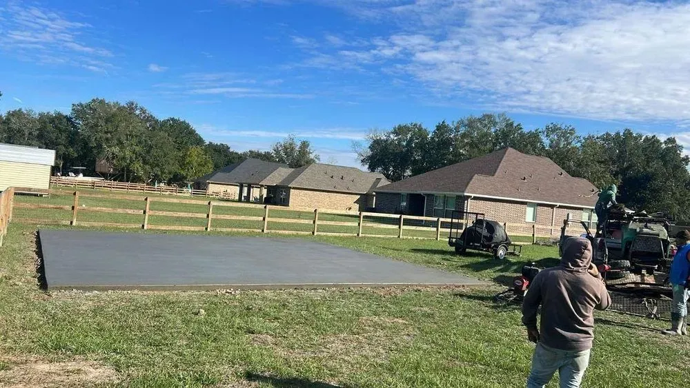 Newly poured concrete slab in a grassy field, flanked by a wooden fence and houses under a blue sky.