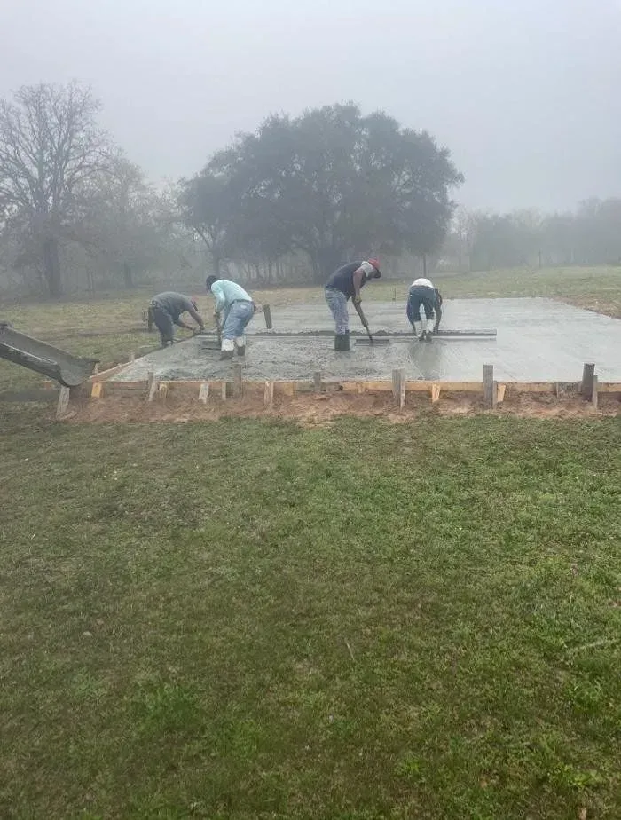 Workers leveling wet concrete on a framed slab in a grassy field on a foggy day.