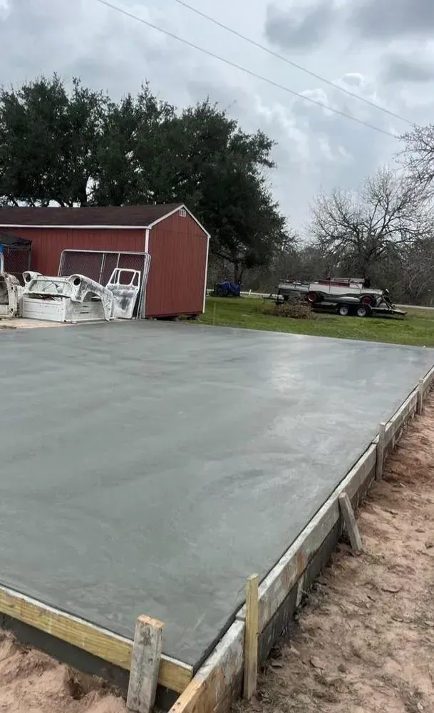 Newly poured concrete slab with wooden formwork, in a rural setting, overcast sky.