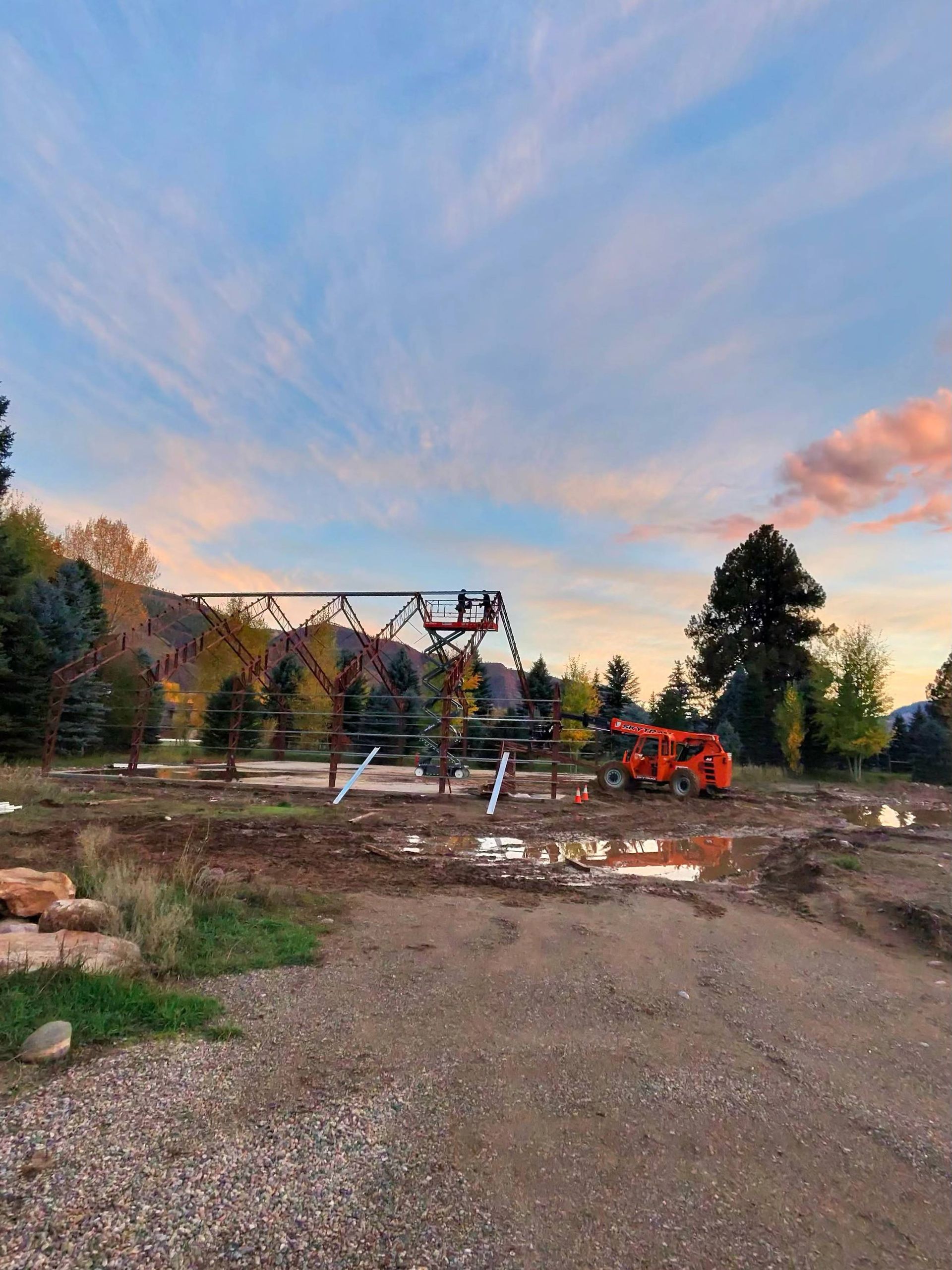Construction site with metal framework against a sunset sky; an orange lift truck is parked nearby.