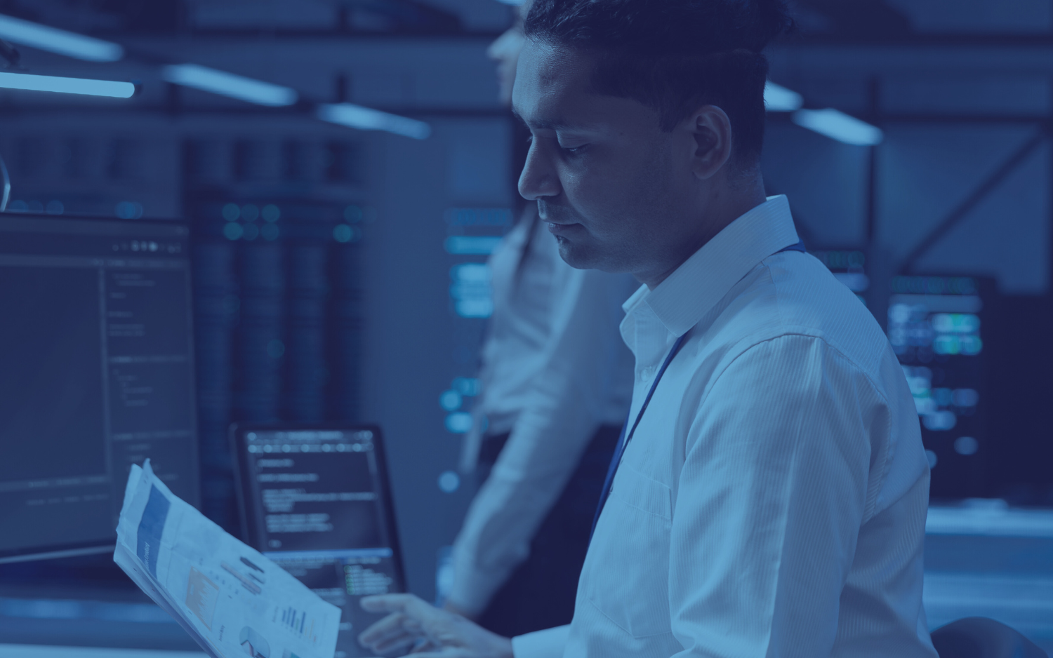 Man in a white shirt examines documents, working in a tech-filled control room.  Blue lighting.