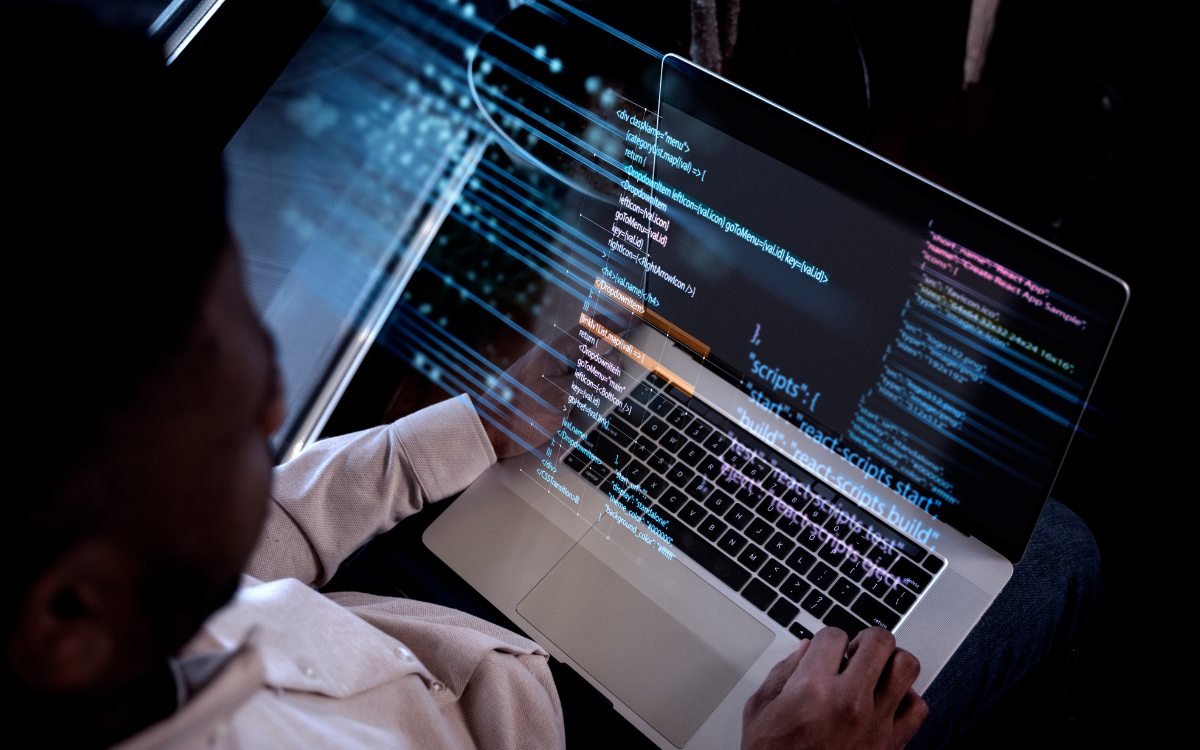 Man working on a laptop, code projected on the screen. Indoors, low lighting.