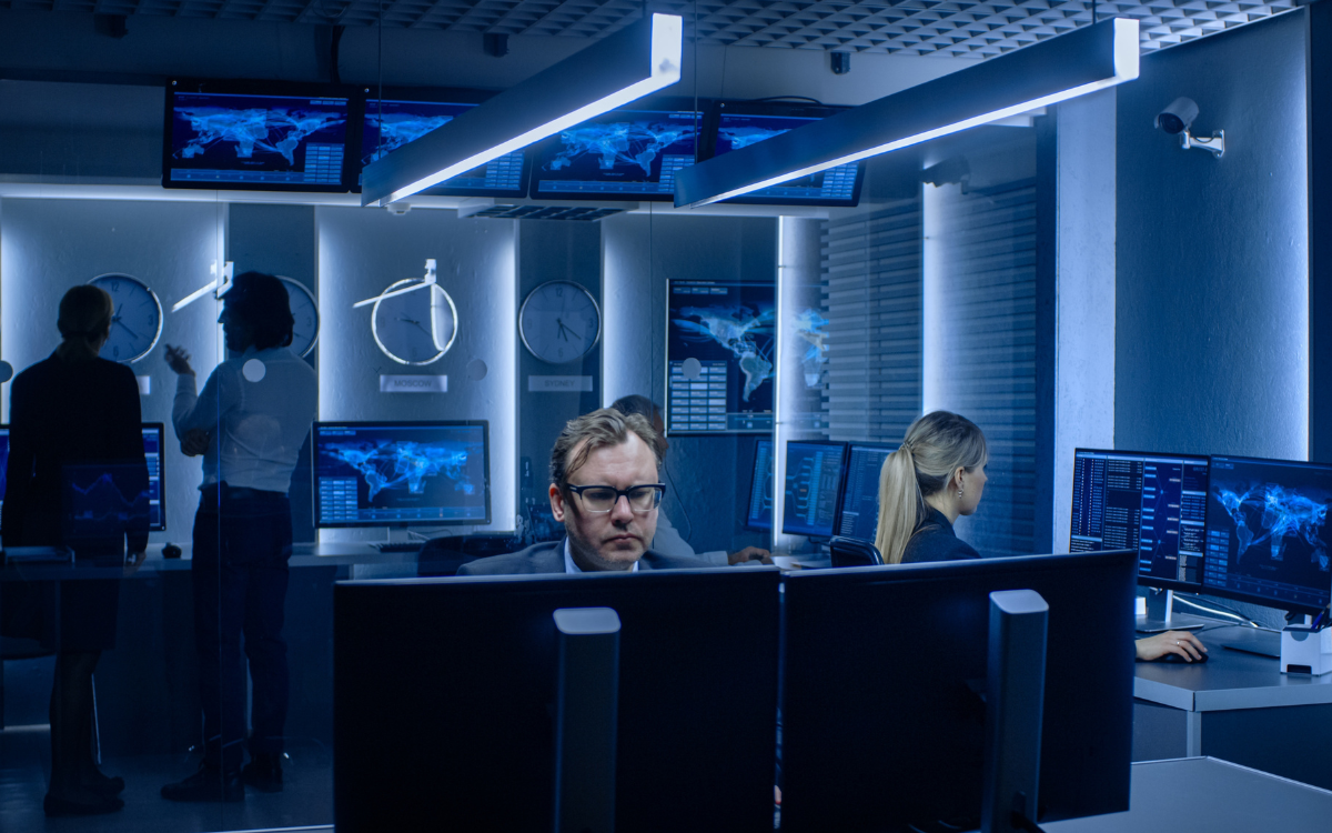 People in a modern control room monitor data on multiple screens. Blue lighting illuminates the scene.