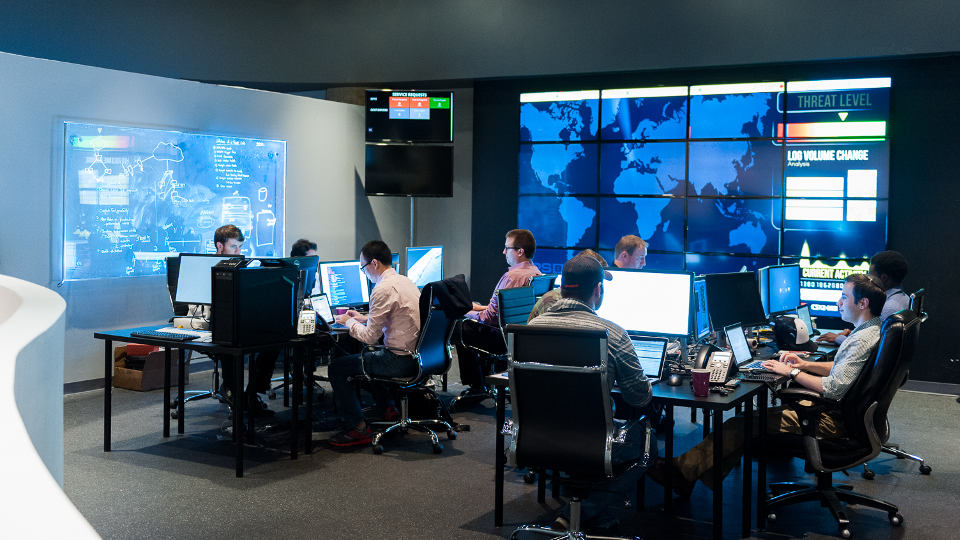 People working in a high-tech control room with multiple computer screens displaying data and world maps. Dim lighting.
