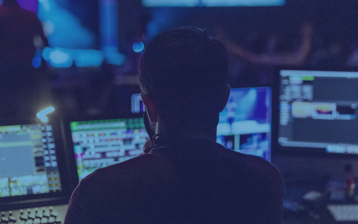 Person facing multiple computer screens in a dark control room, monitoring video feeds.