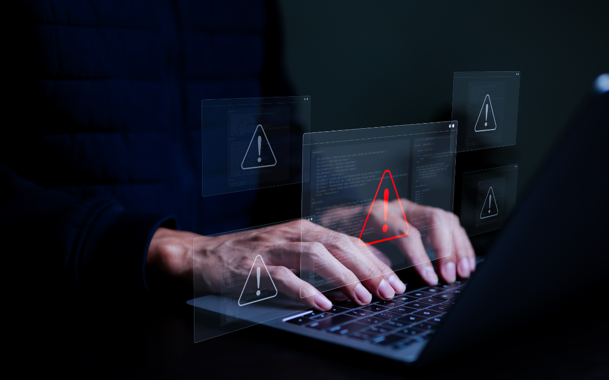 Person typing on a laptop, overlaid with digital warning symbols in a dark setting.