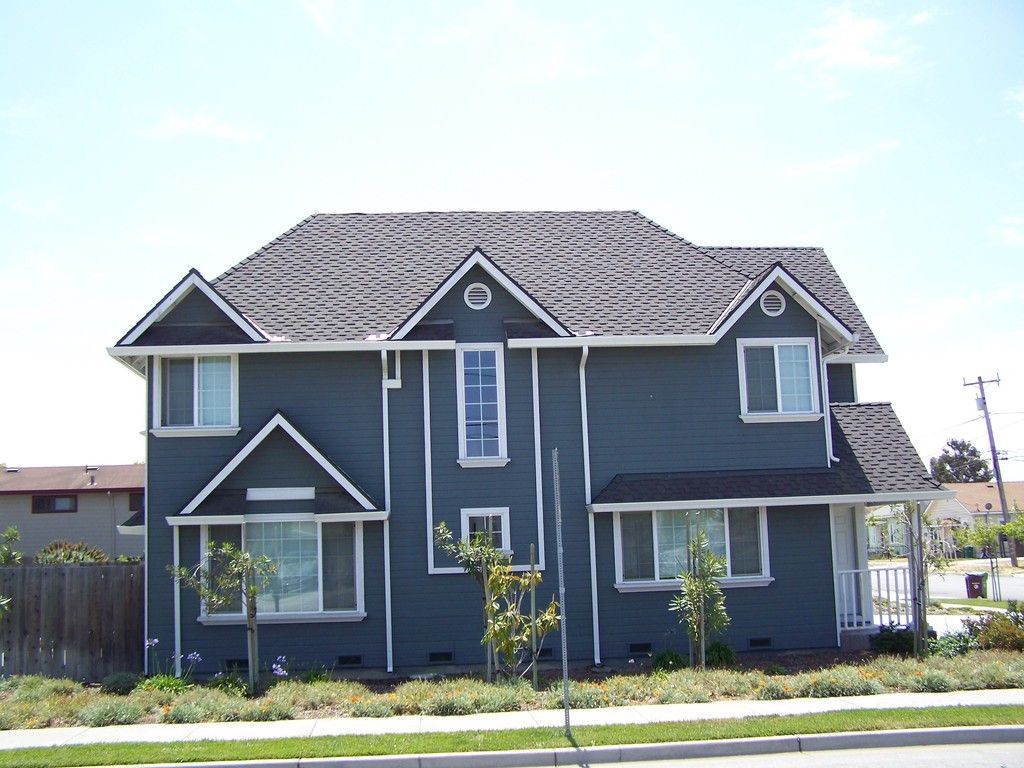Two-story blue house with white trim. Windows, gabled roof, and small front yard with shrubs.