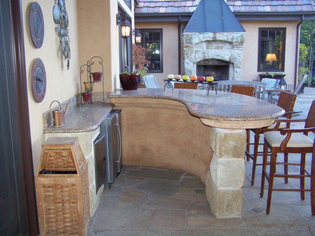 Outdoor bar with granite countertop, stone base, and built-in appliances; chairs nearby, fireplace in background.