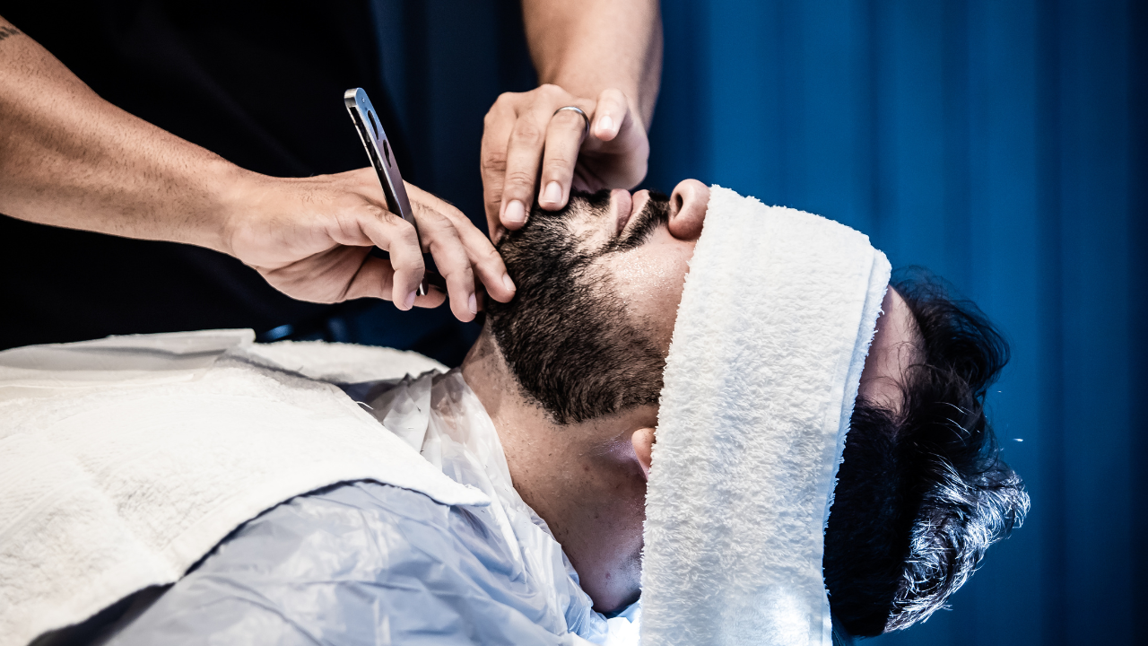 Barber shaving a customer's beard with a straight razor. White towel on face, blue backdrop.