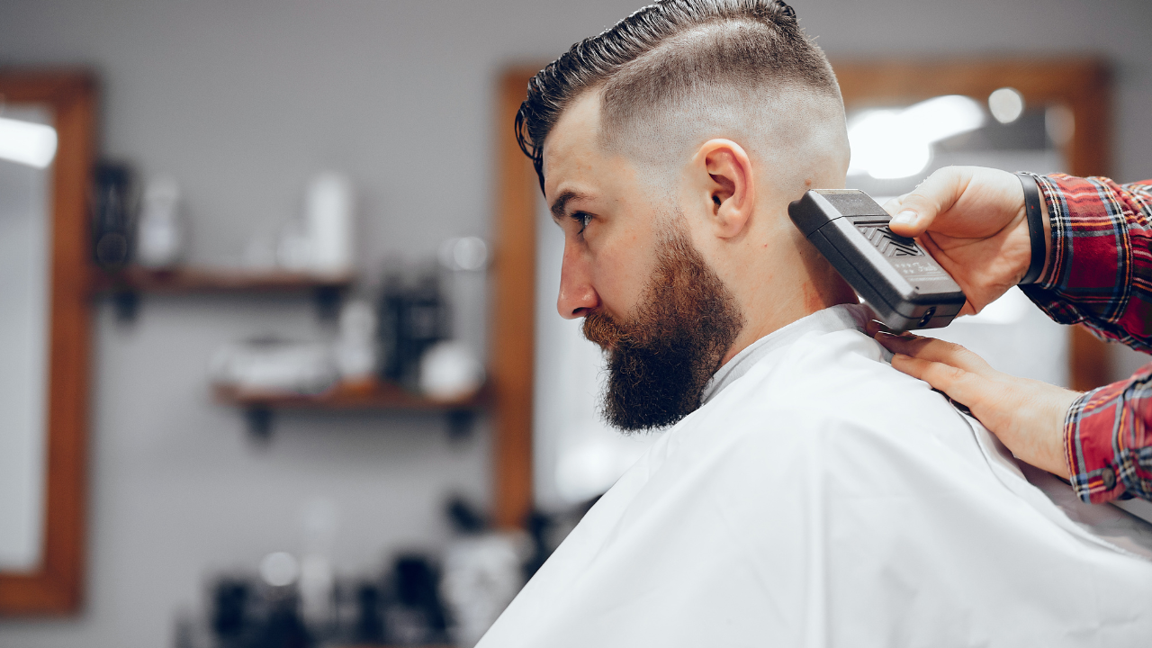 Barber trimming a man's hair with electric clippers in a barbershop. The man has a beard and is wearing a salon cape.