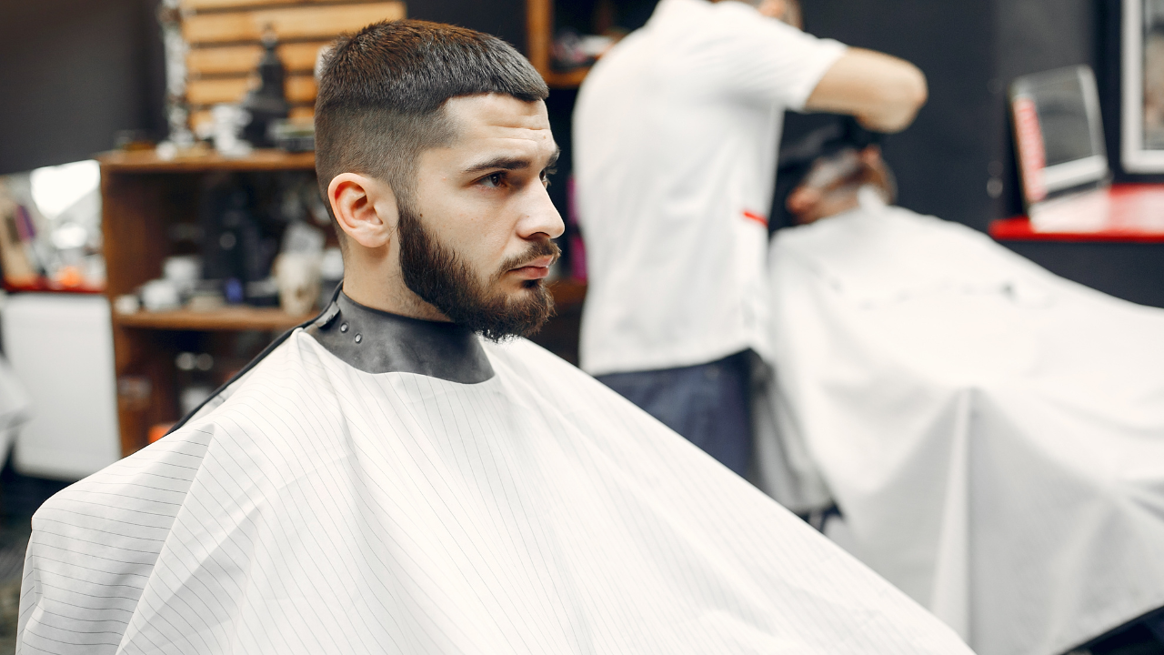 Man with a beard in barber chair, wearing a cape; another person blurred in the background.