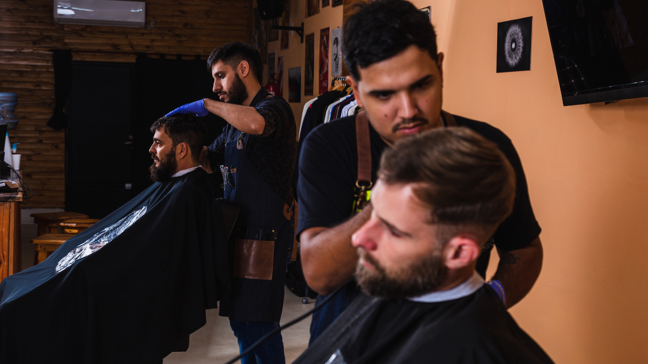 Two barbers give haircuts in a barbershop. One shaves a client's hair, while another uses clippers on a different customer.