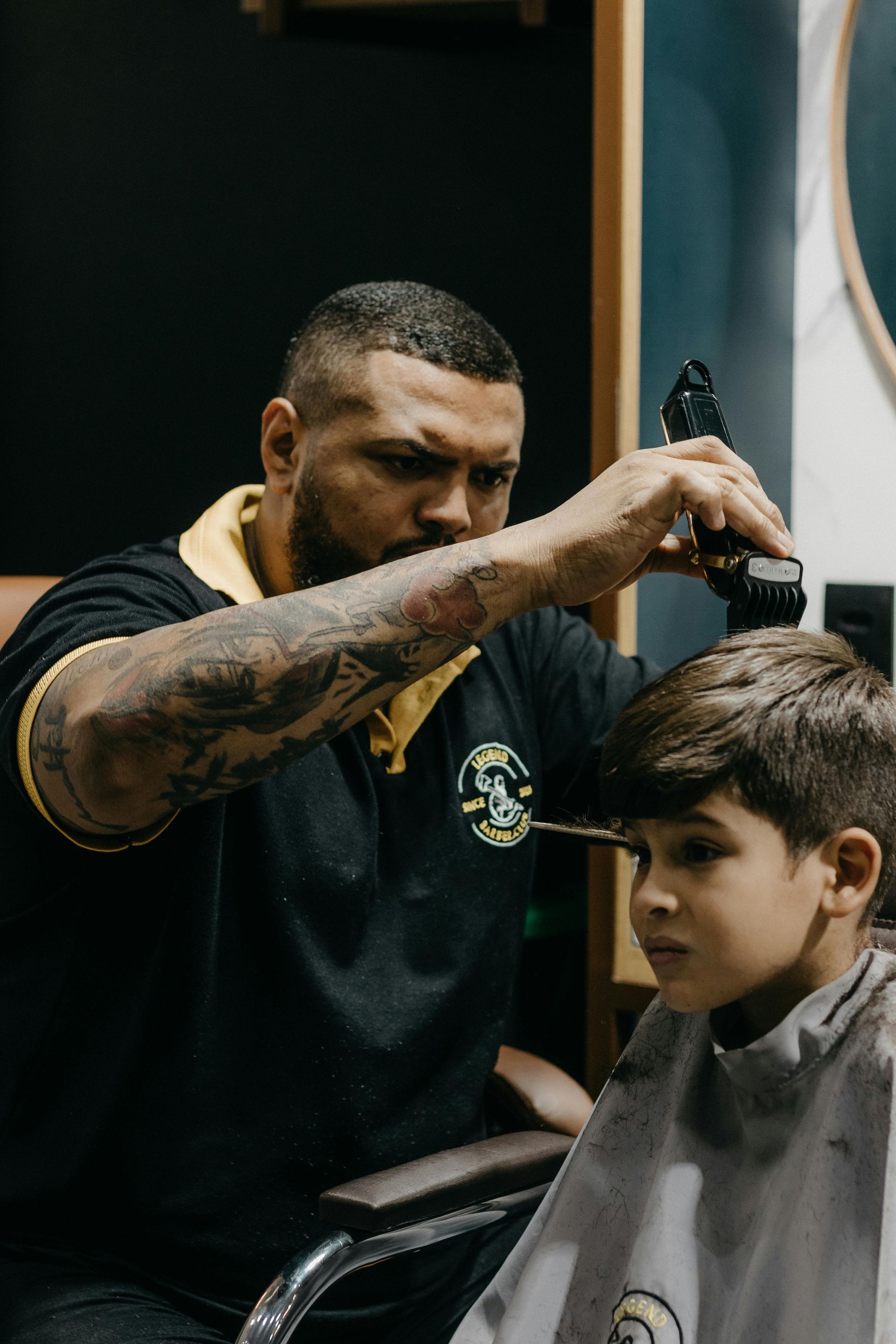 Barber cutting a client's hair with electric clippers. The client wears a striped cape. Low-lit interior.