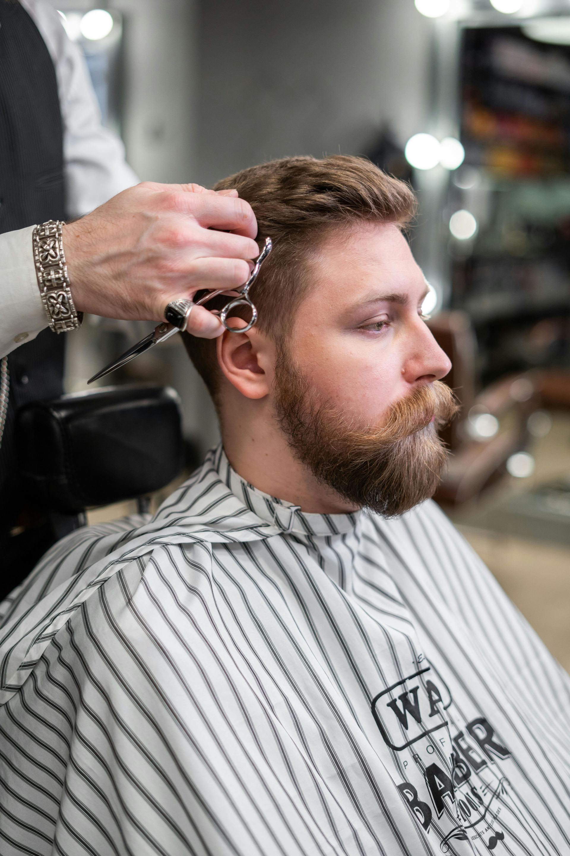Barber cutting a man's hair in a barber shop. Man has a beard, wears a striped cape.