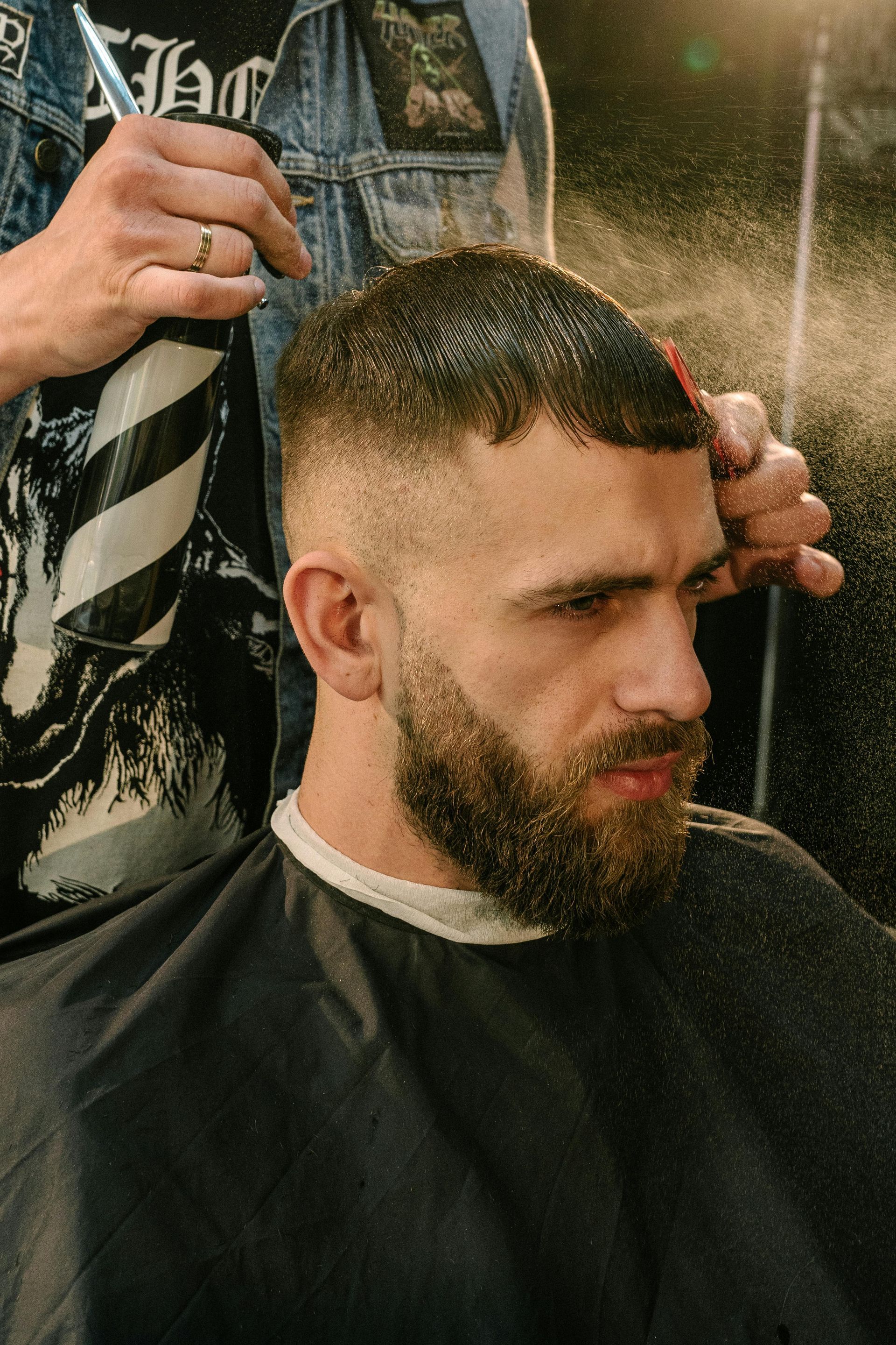 Barber spraying man's hair during a fade haircut, outdoors. Man has a beard, wearing a black cape.