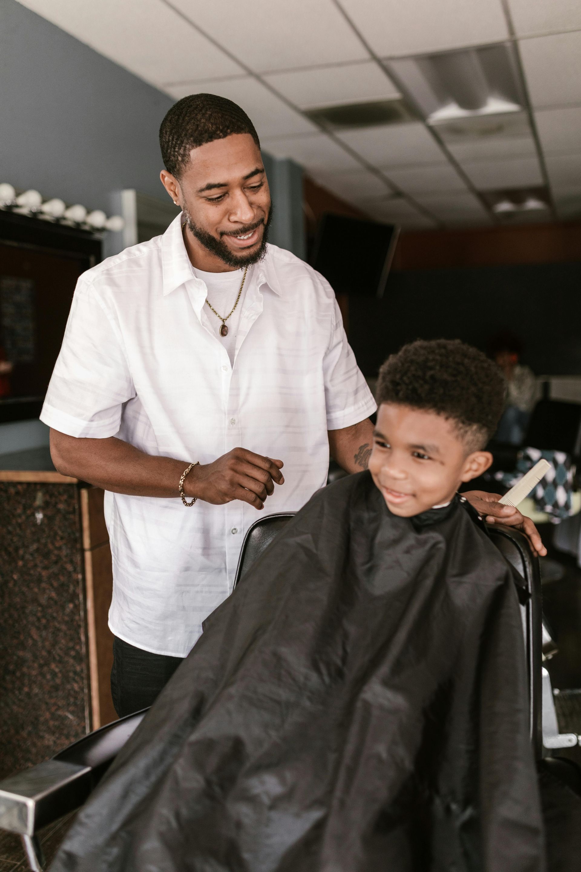Barber cutting a client's hair in a barbershop. The client sits in a chair wearing a cape. The barber has tattoos.