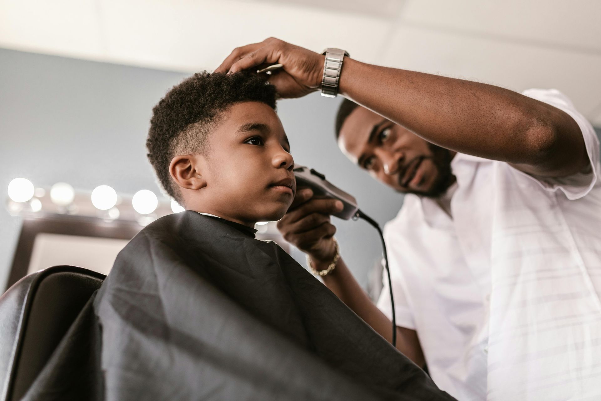 Barber cutting a child's hair in a salon. The child is seated wearing a black cape, while the barber uses clippers.