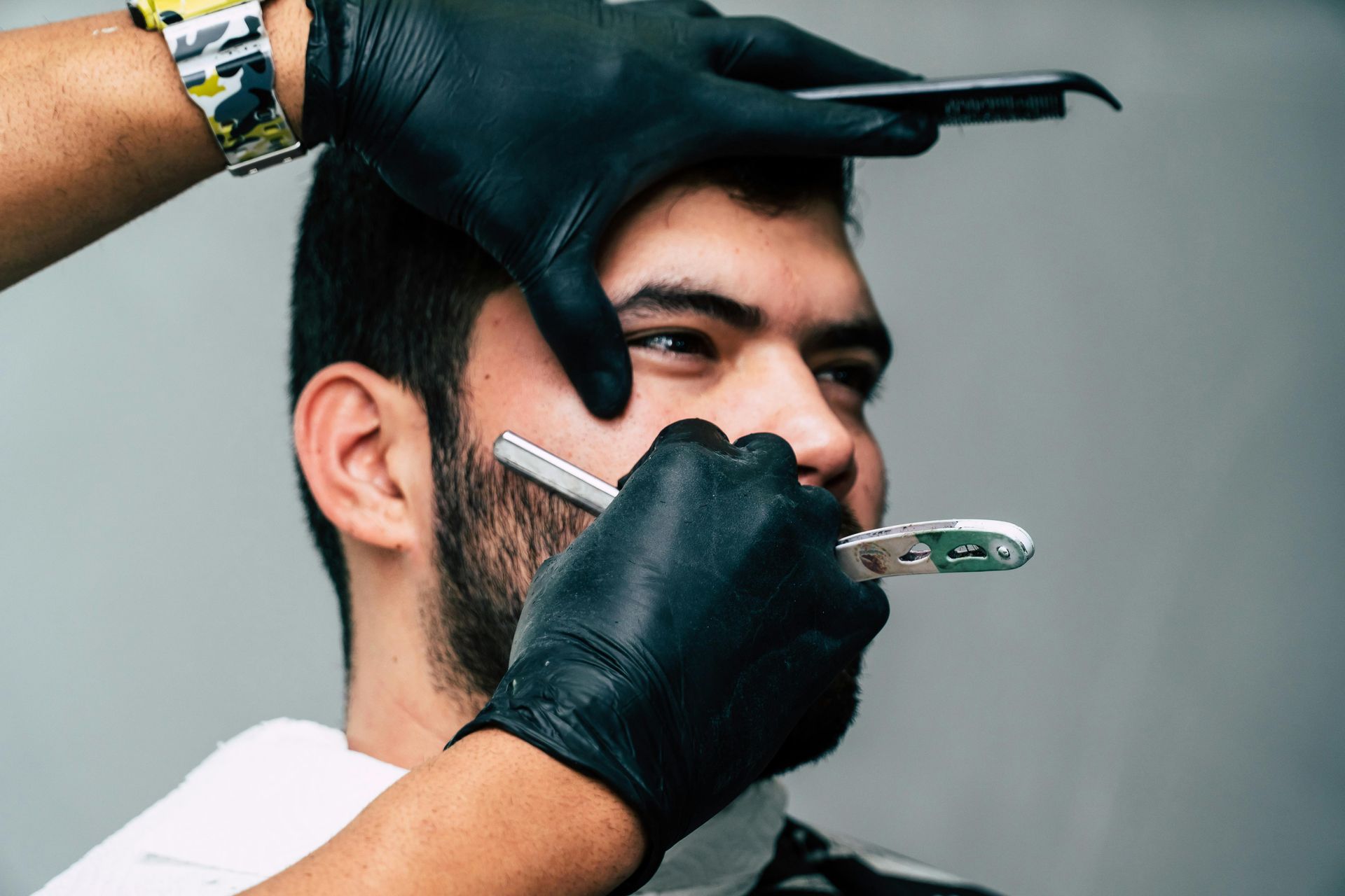 Barber shaves a client's beard with a straight razor; close-up of face; black gloves.