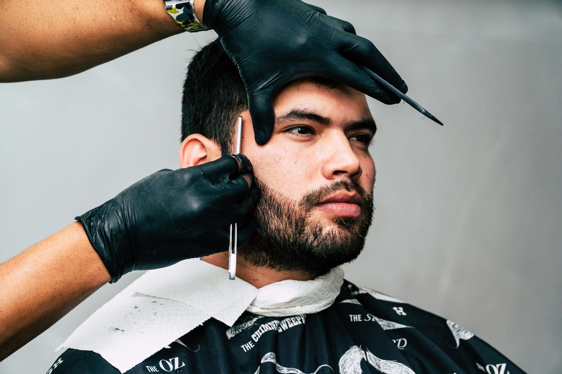 Barber using a straight razor to shape a client's beard. Client wears a cape and has a focused expression.