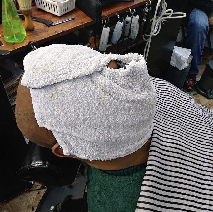 A person in a barber’s chair with a white towel wrapped around their head, positioned for a service in a salon.