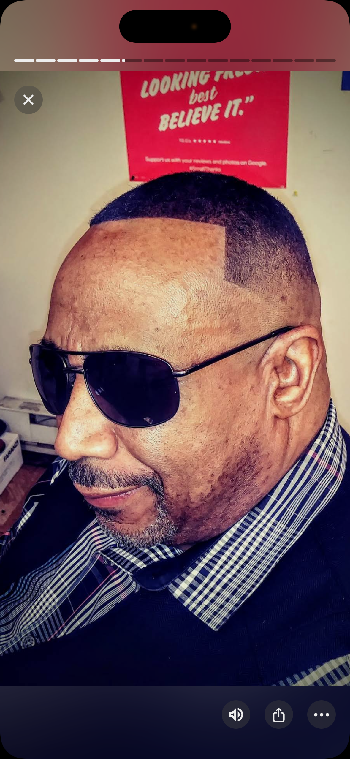 A person in sunglasses and a patterned collared shirt, positioned in front of a red sign with white text.