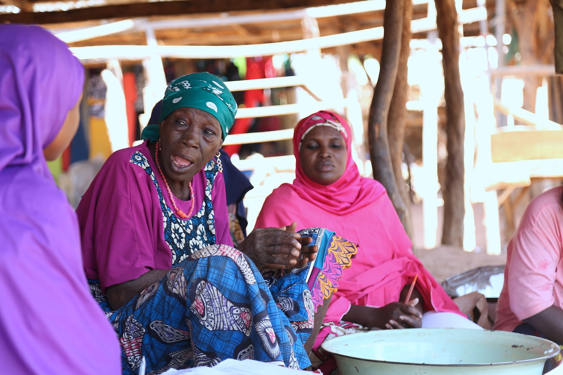 Three women in colorful headscarves sit and converse outdoors.