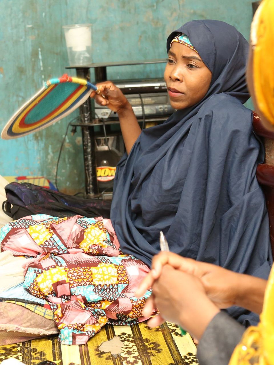 Woman in blue hijab fans herself, speaking; colorful fabric and items in room.