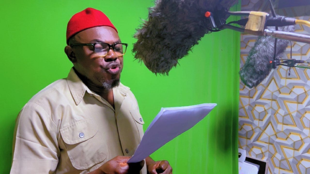 Man in red hat recording audio, reading from papers near a green screen and microphone.