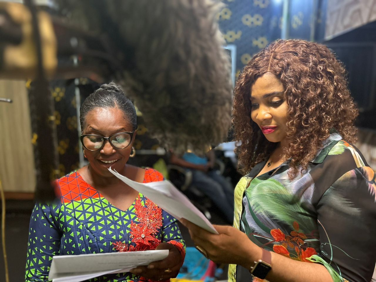 Two women in patterned dresses read papers; one smiles, the other looks down; indoor setting.